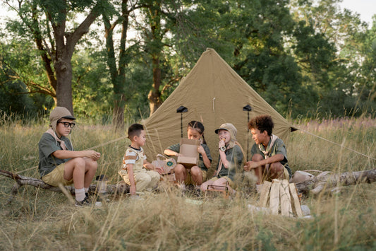 Five children in scout-style outfits sit together in a grassy meadow in front of a canvas tent, sharing snacks and opening a box during a camping adventure surrounded by trees.